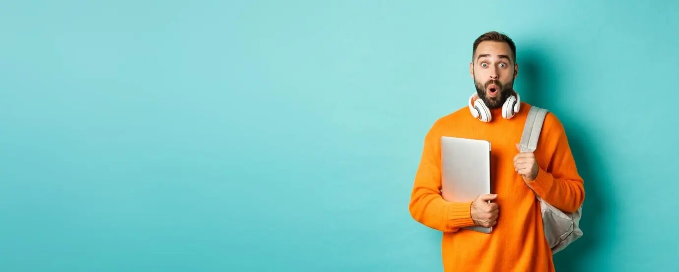 Счастливый мужчина с рюкзаком и наушниками держит ноутбук, улыбается, выглядит удивлённым и стоит над tu.