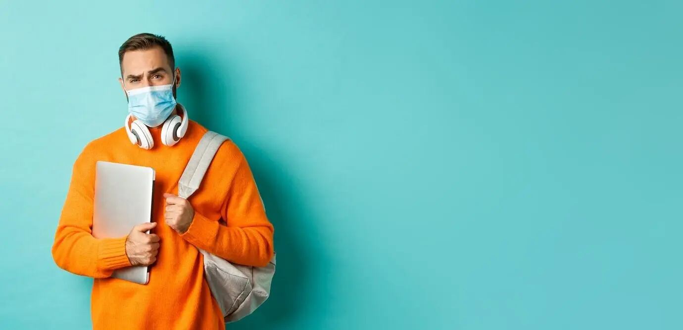 Красивый мужчина европеоидной внешности в наушниках и с рюкзаком, в медицинской маске, держит ноутбук и смотрит.